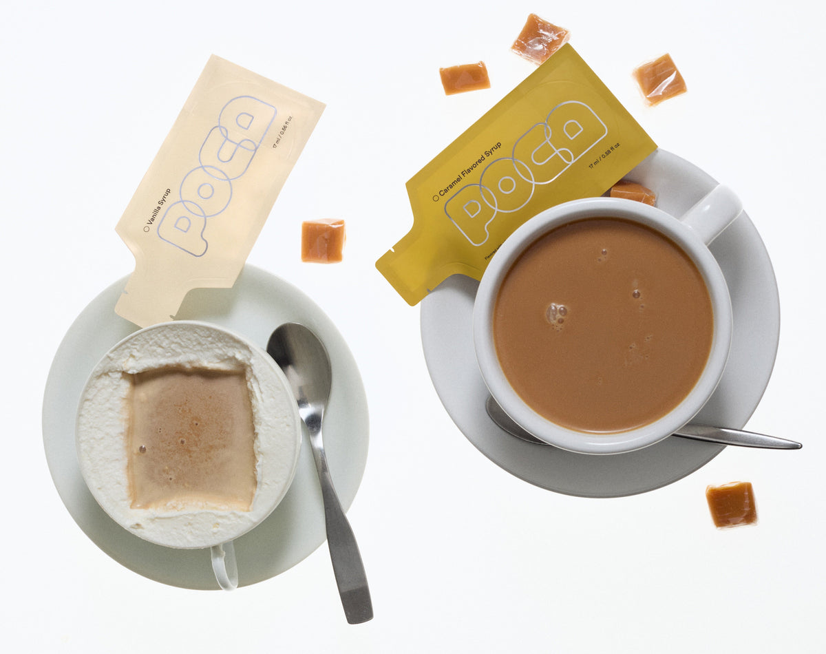 Two cups of coffee with milk foam along the edge and two poca packets on the side.