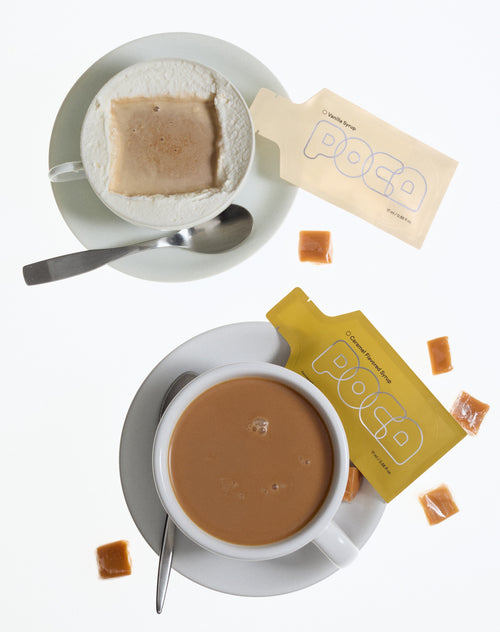 Two cups of coffee with milk foam along the edge and two poca packets on the side.